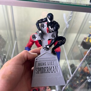 May include: A black and white action figure of Spider-Man is posed on a gray tombstone with the inscription "Here Lies Spiderman".