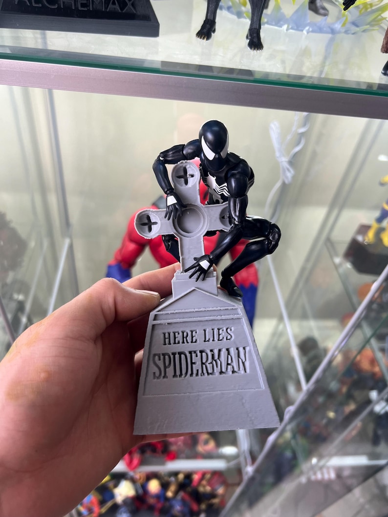 May include: A black and white action figure of Spider-Man is posed on a gray tombstone with the inscription "Here Lies Spiderman".