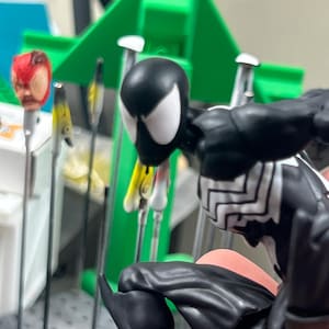 May include: A black and white Spider-Man action figure with a detailed design. The figure is posed in a crouching position. In the background, there are other Spider-Man heads and a green structure.