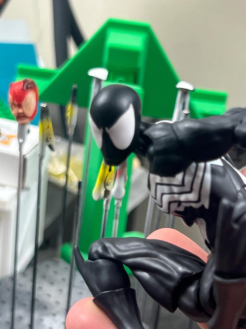 May include: A black and white Spider-Man action figure with a detailed design. The figure is posed in a crouching position. In the background, there are other Spider-Man heads and a green structure.
