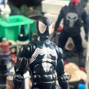May include: A black Spider-Man action figure with white spider-web detailing on the chest and back. The figure has white eye shapes and is posed in front of a blurred background with other action figures and art supplies.