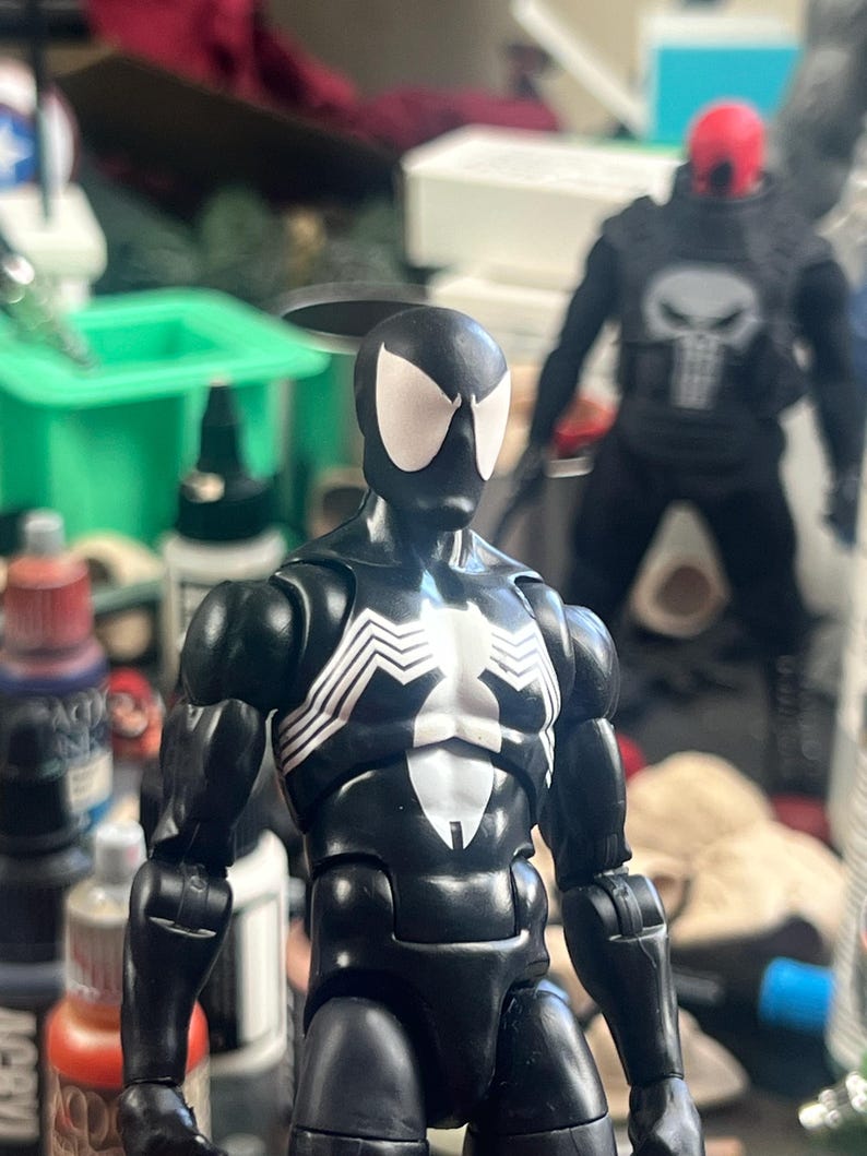 May include: A black Spider-Man action figure with white spider-web detailing on the chest and back. The figure has white eye shapes and is posed in front of a blurred background with other action figures and art supplies.