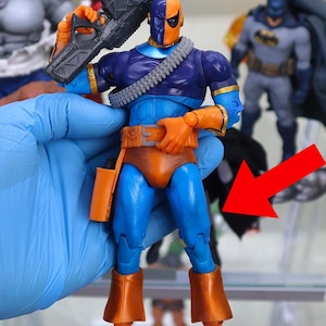 Puede incluir: Figura de acción de Deathstroke con un esquema de color azul y naranja, sosteniendo un arma negra. La figura tiene un diseño detallado, incluyendo un cinturón de herramientas y una bandolera. Otras figuras de superhéroes son visibles en el fondo.