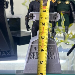 May include: Black Panther action figure standing on a gray base with the words "HE SPIES MAN" printed on it. The figure is 7 inches tall and is being measured with a yellow tape measure.