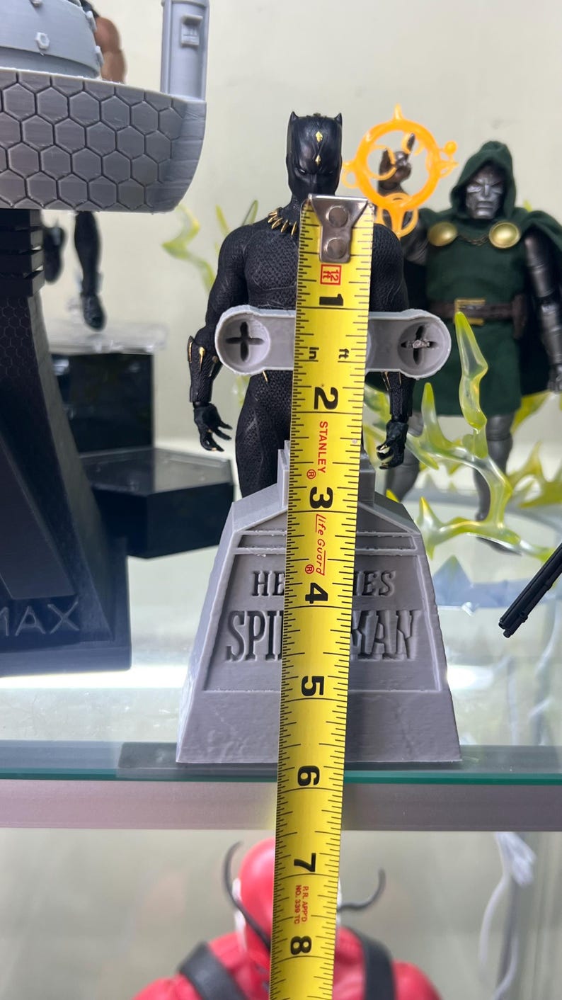 May include: Black Panther action figure standing on a gray base with the words "HE SPIES MAN" printed on it. The figure is 7 inches tall and is being measured with a yellow tape measure.
