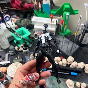 May include: A black and white Spider-Man action figure with a detailed design, held in front of a cluttered workspace. Other action figures and modeling supplies are visible in the background.
