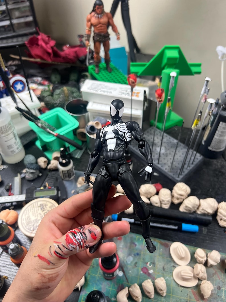 May include: A black and white Spider-Man action figure with a detailed design, held in front of a cluttered workspace. Other action figures and modeling supplies are visible in the background.