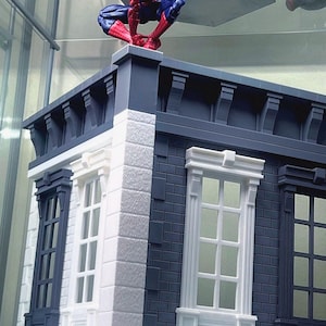 Puede incluir: Figura de acción detallada de Spider-Man en cuclillas sobre una fachada de edificio en miniatura. El edificio presenta ladrillos blancos y azul oscuro, ventanas y molduras decorativas. La figura es roja y azul.