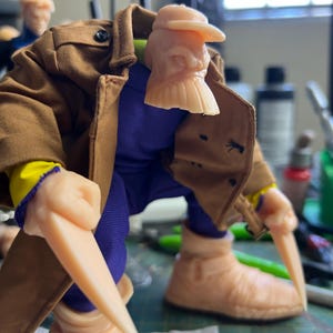 Puede incluir: Figura de acción detallada de un personaje con gabardina marrón, camisa morada y botas color canela. La figura tiene cuchillas color canela que se extienden de sus manos y una cabeza esculpida con sombrero. La figura está posada sobre una superficie verde.