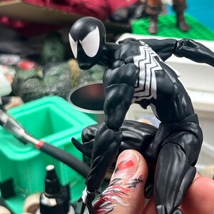 May include: A black and white superhero action figure in a dynamic pose. The figure has a black suit with white spider-web detailing on the chest and back. The figure's head is black with white eye shapes. The figure is articulated and appears to be in mid-action.