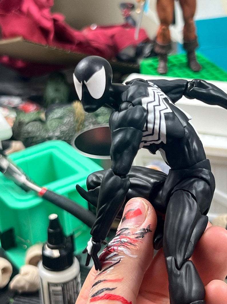 May include: A black and white superhero action figure in a dynamic pose. The figure has a black suit with white spider-web detailing on the chest and back. The figure's head is black with white eye shapes. The figure is articulated and appears to be in mid-action.