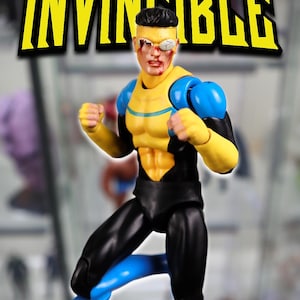 Könnte beinhalten: Eine Invincible-Actionfigur in einem gelb-blauen Anzug mit schwarzen Akzenten. Die Figur hat ein kampfgezeichnetes Gesicht mit einer weißen Augenklappe und roten Blutflecken. Das Wort INVINCIBLE steht in großen gelben Buchstaben über der Figur.