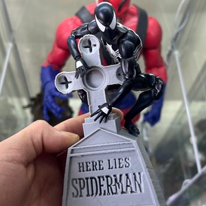 May include: A black and white action figure of Spider-Man kneeling on a gray tombstone with the words "Here Lies Spiderman" inscribed on it. The figure is posed as if it is being buried.
