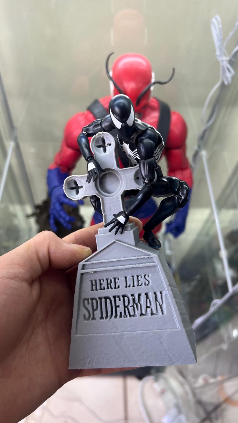 May include: A black and white action figure of Spider-Man kneeling on a gray tombstone with the words "Here Lies Spiderman" inscribed on it. The figure is posed as if it is being buried.