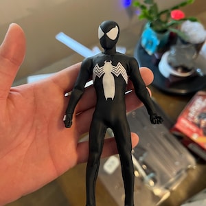 Puede incluir: Figura de acción negra de Spider-Man con un traje negro y un emblema de araña blanco. La figura tiene formas de ojos blancos en la máscara. La figura se sostiene en una mano. Es un objeto de colección.
