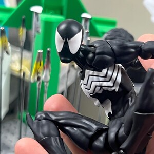 May include: A black and white superhero action figure in a dynamic pose. The figure has a black suit with white spider-webbing details on the chest and back. The figure's head is black with white eye shapes.
