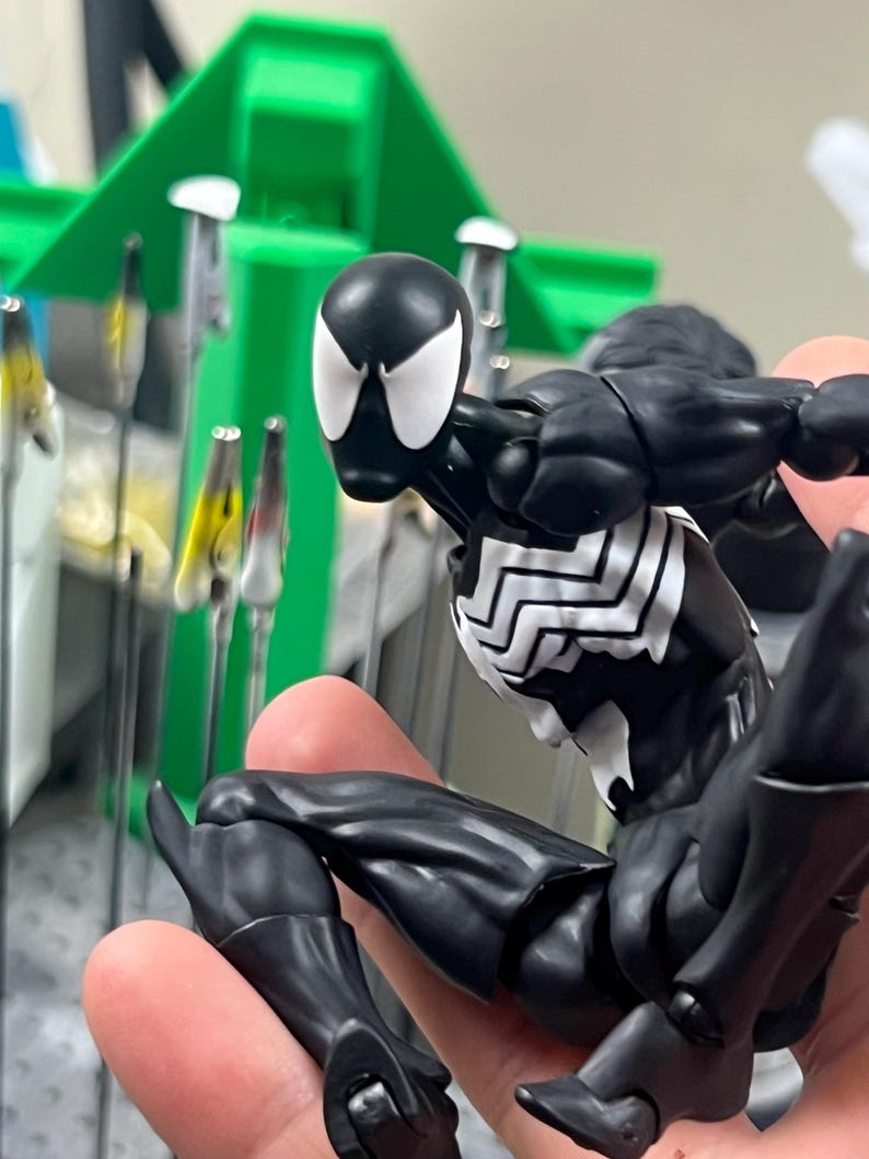May include: A black and white superhero action figure in a dynamic pose. The figure has a black suit with white spider-webbing details on the chest and back. The figure's head is black with white eye shapes.