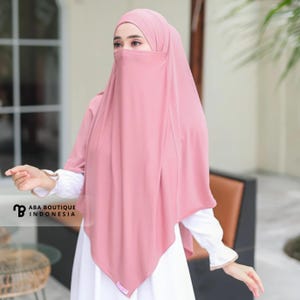 Könnte beinhalten: Ein rosa Hijab- und Niqab-Set. Der Hijab bedeckt Kopf und Schultern, während der Niqab das untere Gesicht bedeckt. Das Outfit wird mit einem weißen Kleid mit Rüschenärmeln kombiniert. Das Logo von ABA Boutique Indonesia ist sichtbar.