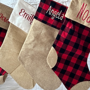 May include: Four personalized Christmas stockings with a red and black buffalo plaid design. The stockings are made of burlap and have a white cuff with the names "Dad", "Emily", "Angela", and "Noah" embroidered in red.