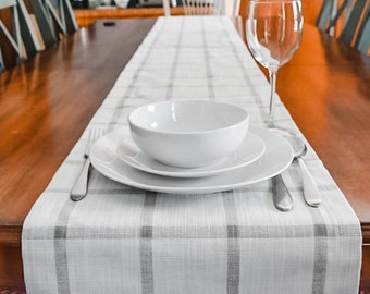 May include: A white ceramic bowl and two white ceramic plates are set on a gray and white plaid table runner. The table runner is on a wooden table.