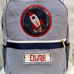 May include: A blue and white striped backpack with a rocket ship applique and a name tag that says "Elijah".