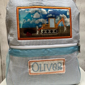 Pode incluir: Uma mochila azul e branca listrada com uma janela de plástico transparente que mostra uma escavadeira amarela. A mochila tem uma borda azul e uma etiqueta com o nome "Oliver".