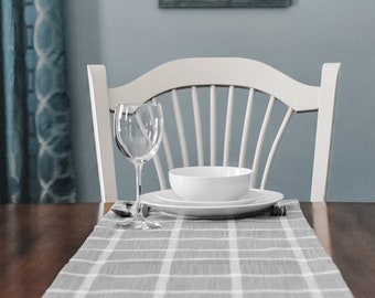 May include: A white chair with a gray and white checkered table runner, a white bowl, a white plate, and a clear wine glass.
