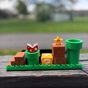 May include: A 3D-printed diorama featuring elements from the Super Mario video game series. It includes a green pipe, a question mark block, a star, and a Piranha Plant. The base has green grass and brown brick platforms.