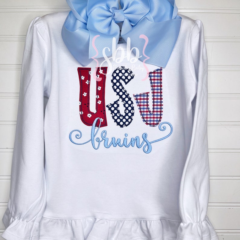 May include: White long-sleeved shirt with a ruffled hem. The shirt features a red, white, and blue applique of the letters "USU" with the word "bruins" embroidered in blue below.