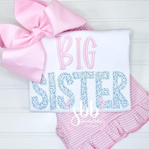May include: A white t-shirt with pink embroidery that says "BIG SISTER" with a floral pattern. The shirt is paired with a pink and white striped skirt with a ruffle.