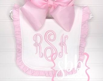 Puede incluir: Babero blanco para bebé con ribete de volantes rosas y un gran lazo rosa. El babero presenta un monograma rosa bordado y está colgado de una percha blanca. Accesorio para bebé.