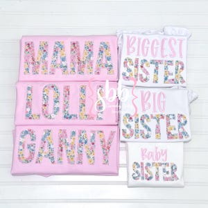 K&ouml;nnte beinhalten: Satz von f&uuml;nf rosa und wei&szlig;en T-Shirts mit Blumenmuster-Schriftzug. Die Shirts tragen die Aufschriften "Mama", "Lolli", "Ganny", "Biggest Sister", "Big Sister" und "Baby Sister".