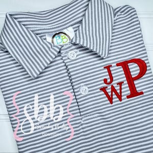 May include: Gray and white striped polo shirt with red embroidered initials "JPW". The shirt has a collared neckline and three white buttons. The text "sbb Swanky Baby boutique" is printed on the shirt in a pink and white design.