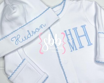 Puede incluir: Conjunto de ropa de bebé blanco con ribetes y bordados en azul claro. El conjunto incluye un gorro con "Hudson" bordado y un body con las iniciales "HHM". El conjunto tiene un borde festoneado.