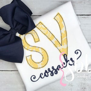 May include: A white t-shirt with a yellow and navy blue monogram "SV" and the text "cossack" in navy blue cursive. The shirt is decorated with a navy blue bow.