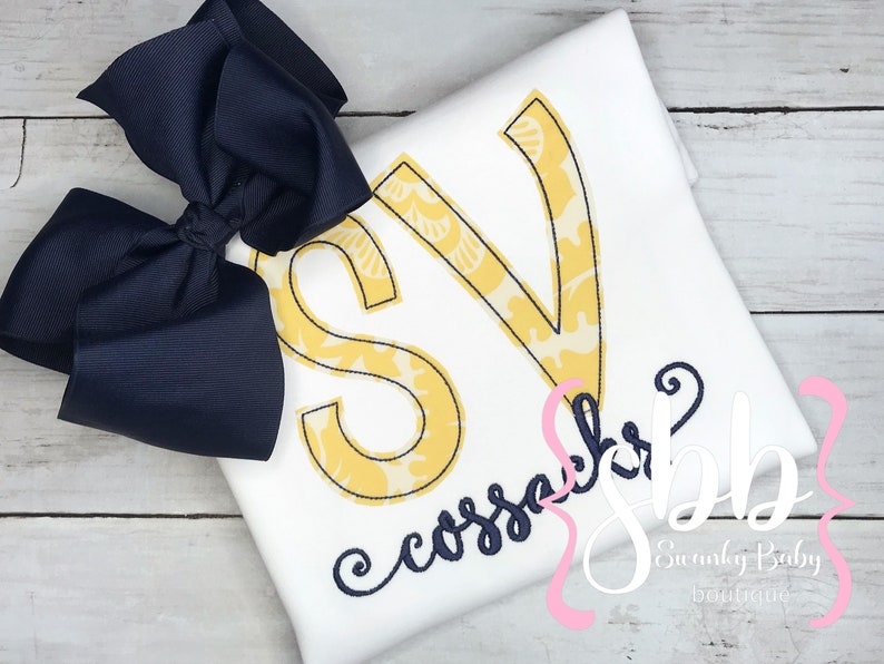 May include: A white t-shirt with a yellow and navy blue monogram "SV" and the text "cossack" in navy blue cursive. The shirt is decorated with a navy blue bow.