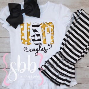 May include: White baby outfit with a black and white striped leggings and a white shirt with a yellow, black, and white "USM Eagles" graphic.