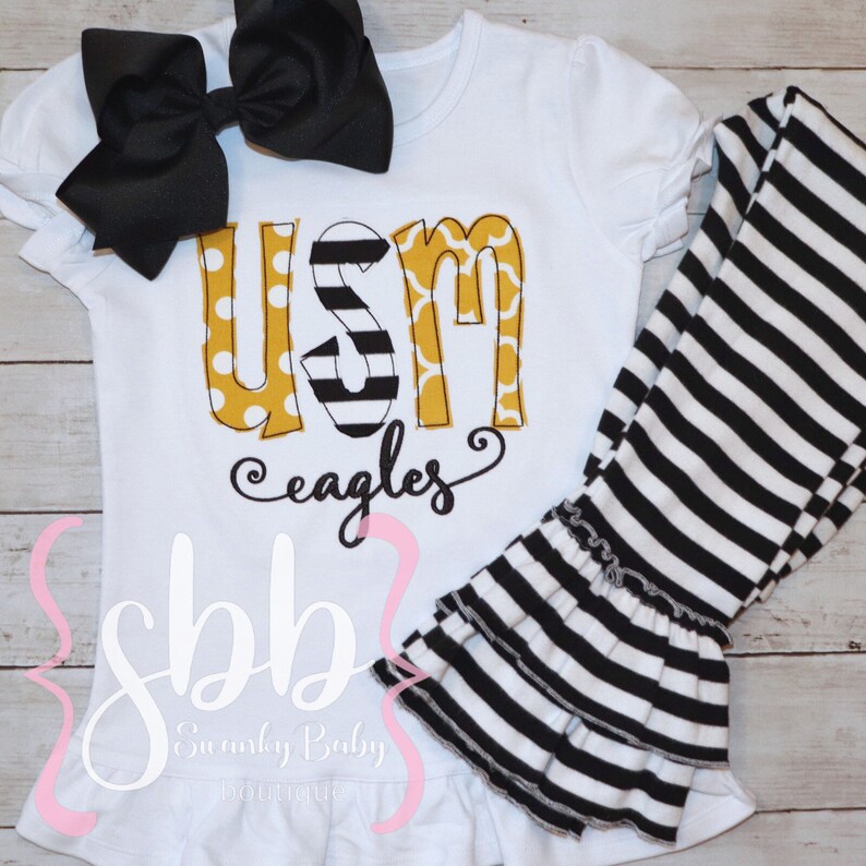 May include: White baby outfit with a black and white striped leggings and a white shirt with a yellow, black, and white "USM Eagles" graphic.
