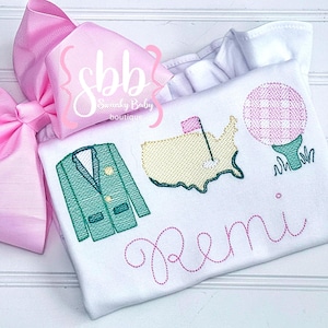 May include: A white onesie with green and pink embroidery. The embroidery includes a golf club, a golf ball, a green jacket, and a map of the United States. The onesie is personalized with the name "Remi".