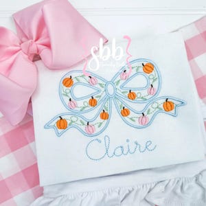 May include: White top with a light blue bow design embroidered with orange and pink pumpkins. The name "Claire" is embroidered in blue script below the bow. A large pink bow is in the upper left corner.