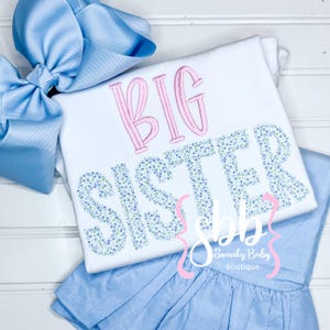May include: White shirt with the words "BIG SISTER" embroidered in pink and blue floral print. The shirt is paired with a light blue skirt.