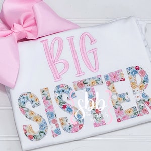 K&ouml;nnte beinhalten: Wei&szlig;er Strampler mit rosa gesticktem Text "BIG SISTER", wobei das Wort "SISTER" ein Blumenmuster hat.