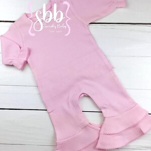 K&ouml;nnte beinhalten: Ein hellrosa Baby-Strampler mit langen &Auml;rmeln und ausgestellten Beinen. Der Strampler hat eine R&uuml;schenborte an den &Auml;rmeln und Beinen. Der Text "sbb Swanky Baby boutique" ist auf der Vorderseite aufgedruckt.