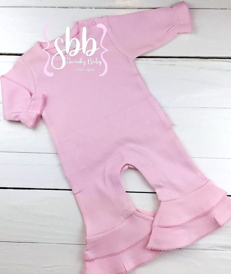 K&ouml;nnte beinhalten: Ein hellrosa Baby-Strampler mit langen &Auml;rmeln und ausgestellten Beinen. Der Strampler hat eine R&uuml;schenborte an den &Auml;rmeln und Beinen. Der Text "sbb Swanky Baby boutique" ist auf der Vorderseite aufgedruckt.