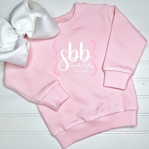 K&ouml;nnte beinhalten: Hellrosa Sweatshirt mit dem Text "sbb Swanky Baby boutique" in wei&szlig;er Schrift und rosa Akzenten. Eine gro&szlig;e wei&szlig;e Schleife befindet sich auf der linken Seite des Sweatshirts. Das Sweatshirt hat gerippte B&uuml;ndchen und einen Saum.