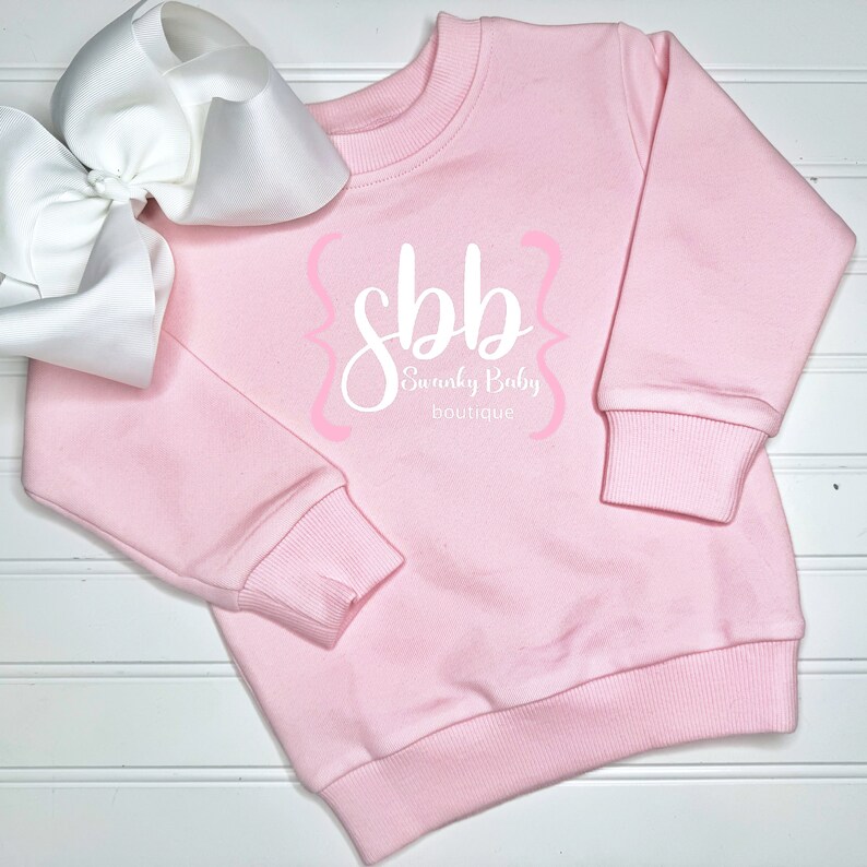 K&ouml;nnte beinhalten: Hellrosa Sweatshirt mit dem Text "sbb Swanky Baby boutique" in wei&szlig;er Schrift und rosa Akzenten. Eine gro&szlig;e wei&szlig;e Schleife befindet sich auf der linken Seite des Sweatshirts. Das Sweatshirt hat gerippte B&uuml;ndchen und einen Saum.