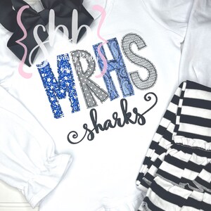May include: White long-sleeved shirt with a ruffled hem, featuring a blue and gray patchwork design that spells out "MRHS" and the text "sharks". The shirt is paired with black and white striped leggings.
