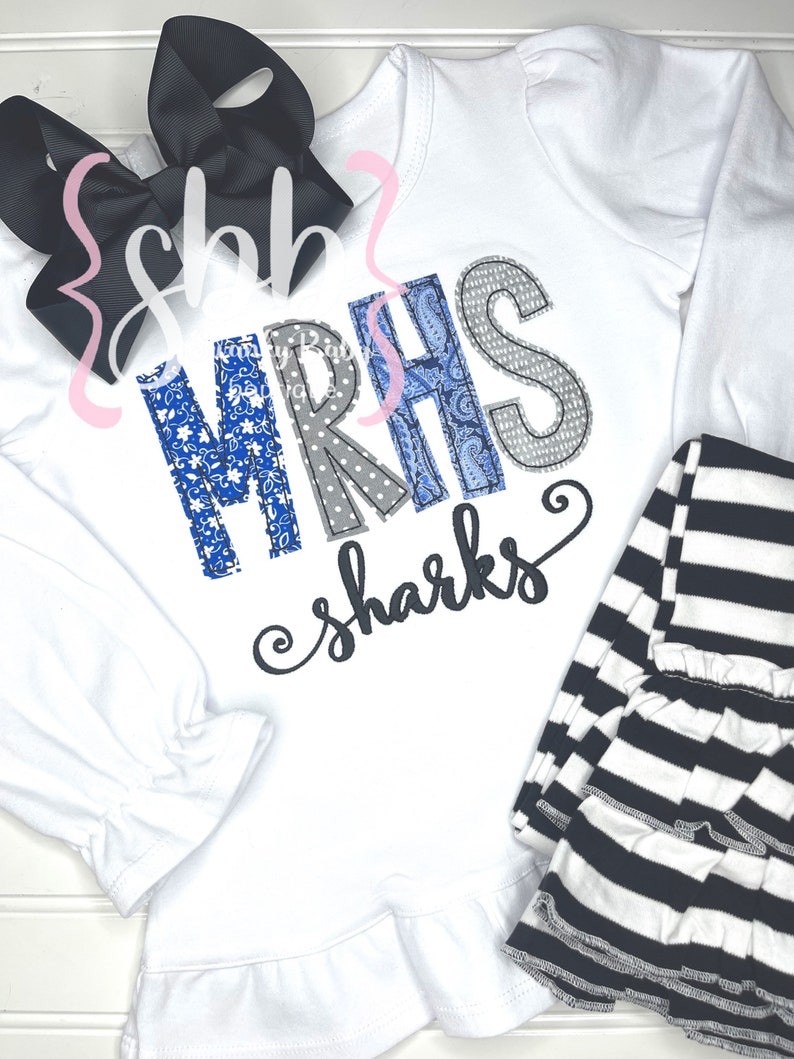 May include: White long-sleeved shirt with a ruffled hem, featuring a blue and gray patchwork design that spells out "MRHS" and the text "sharks". The shirt is paired with black and white striped leggings.
