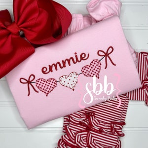 K&ouml;nnte beinhalten: Ein hellrosa Shirt mit dem Namen "emmie" in Rot gestickt, zusammen mit einer Kette aus Herzen. Die Herzen sind mit Mustern verziert und durch rote Schleifen verbunden. Eine rot-wei&szlig; gestreifte Hose und eine gro&szlig;e rote Schleife sind ebenfalls zu sehen.