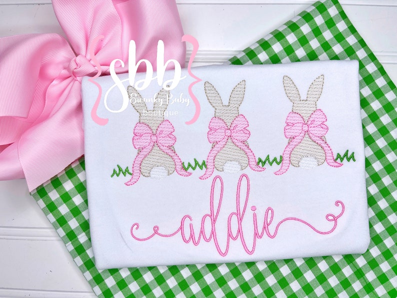 May include: White embroidered t-shirt with three brown bunnies wearing pink bows. The bunnies are standing on a green grass design. The text "sbb" and "Sunny Baby Boutique" are embroidered in the top left corner. The name "addie" is embroidered in pink cursive below the bunnies.